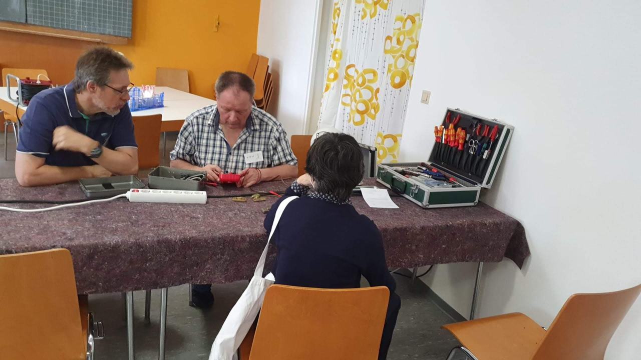 An einem Tisch wird ein Gerät mit dem Multimeter geprüft, daneben steht ein geöffneter Werkzeugkoffer.