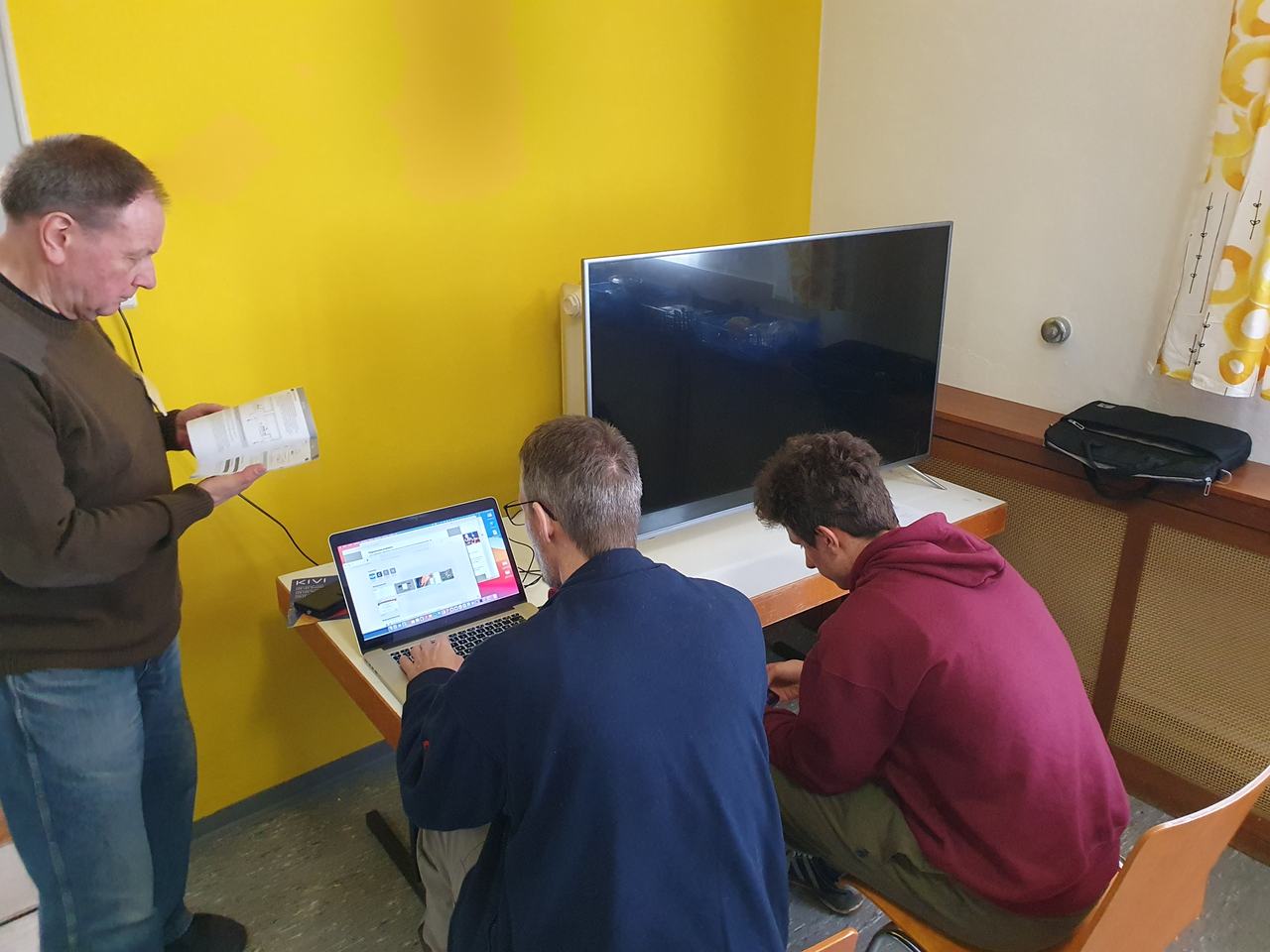 Zwei Helfer richten am Laptop einen Fernseher ein, ein weiterer hält ein ausgebautes Bauteil in den Händen.