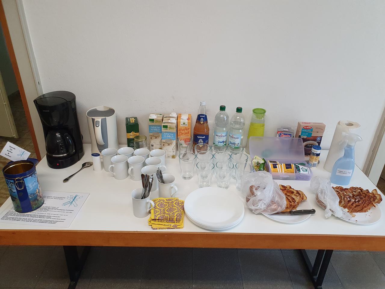 Gedeckter Tisch mit Kaffeemaschine, Tassen, Gläsern, Getränken und zwei Blechkuchen.